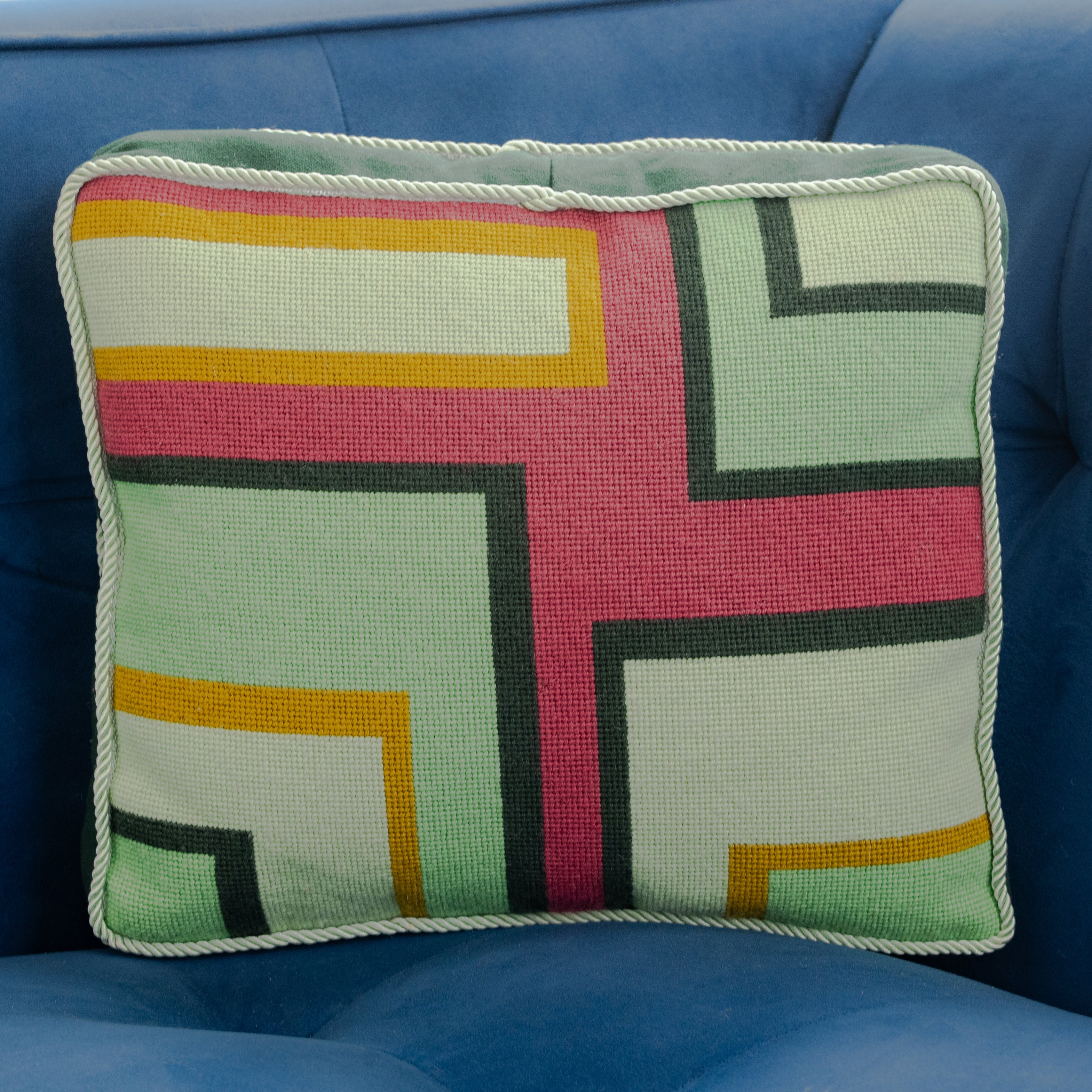 A colorful geometric patterned needlepointed cushion on a blue couch. Designed and sold by Daisy’s Needlepoint Kits.