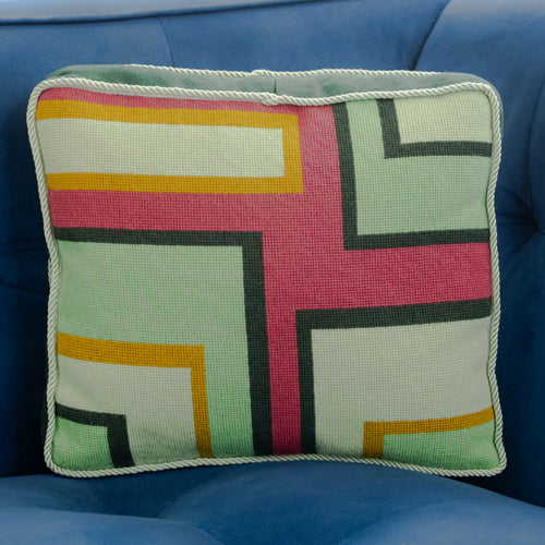 A colorful geometric patterned needlepointed cushion on a blue couch. Designed and sold by Daisy’s Needlepoint Kits.