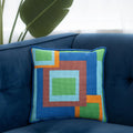 A colorful geometric patterned needlepointed cushion on a blue couch. Designed and sold by Daisy’s Needlepoint Kits.
