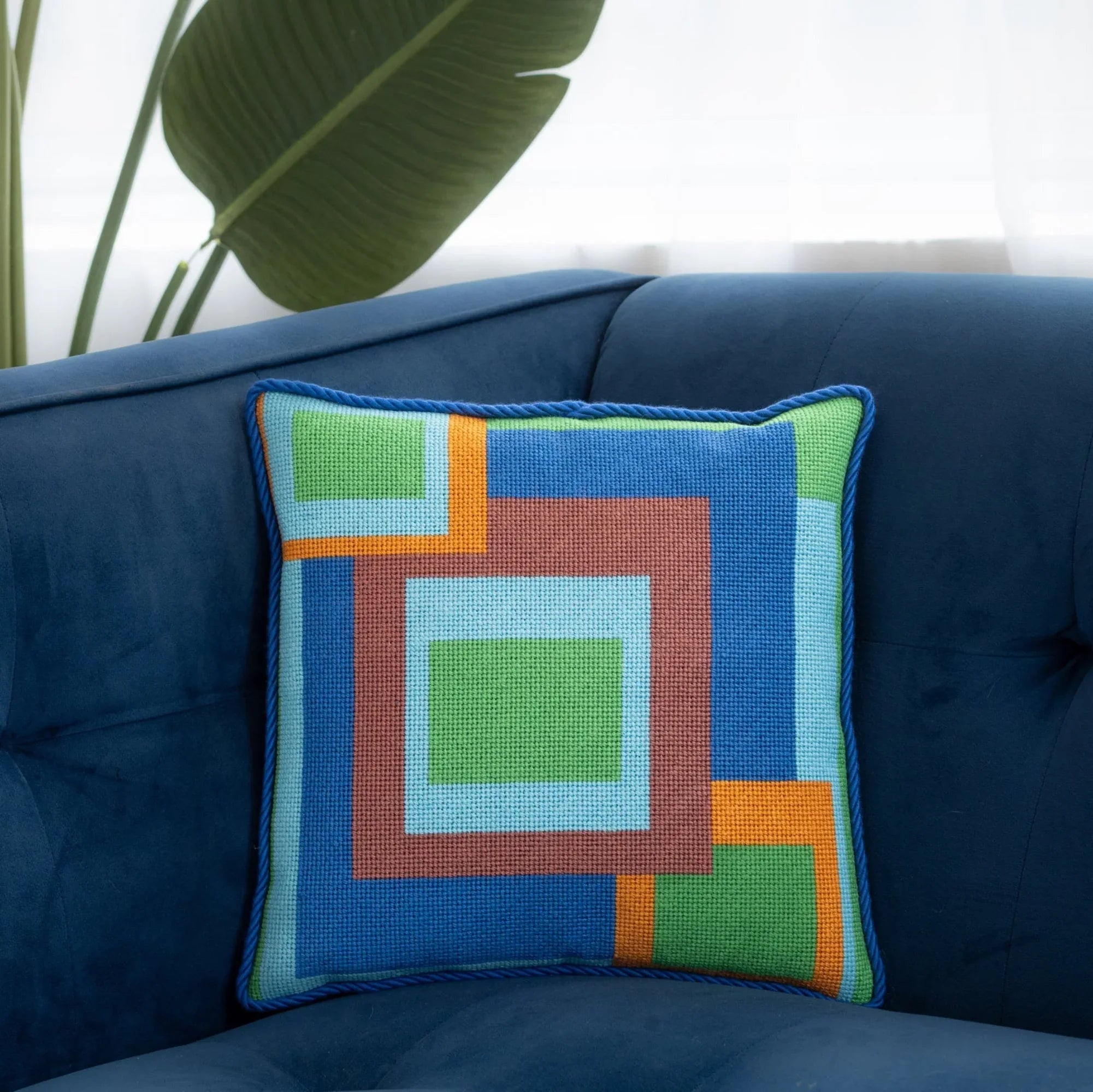 A colorful geometric patterned needlepointed cushion on a blue couch. Designed and sold by Daisy’s Needlepoint Kits.
