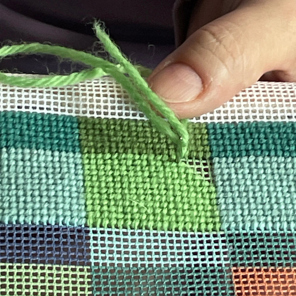 Hand holding green yarn over a cross-stitched needlepoint canvas. Designed and sold by Daisy's Needlepoint Kits.