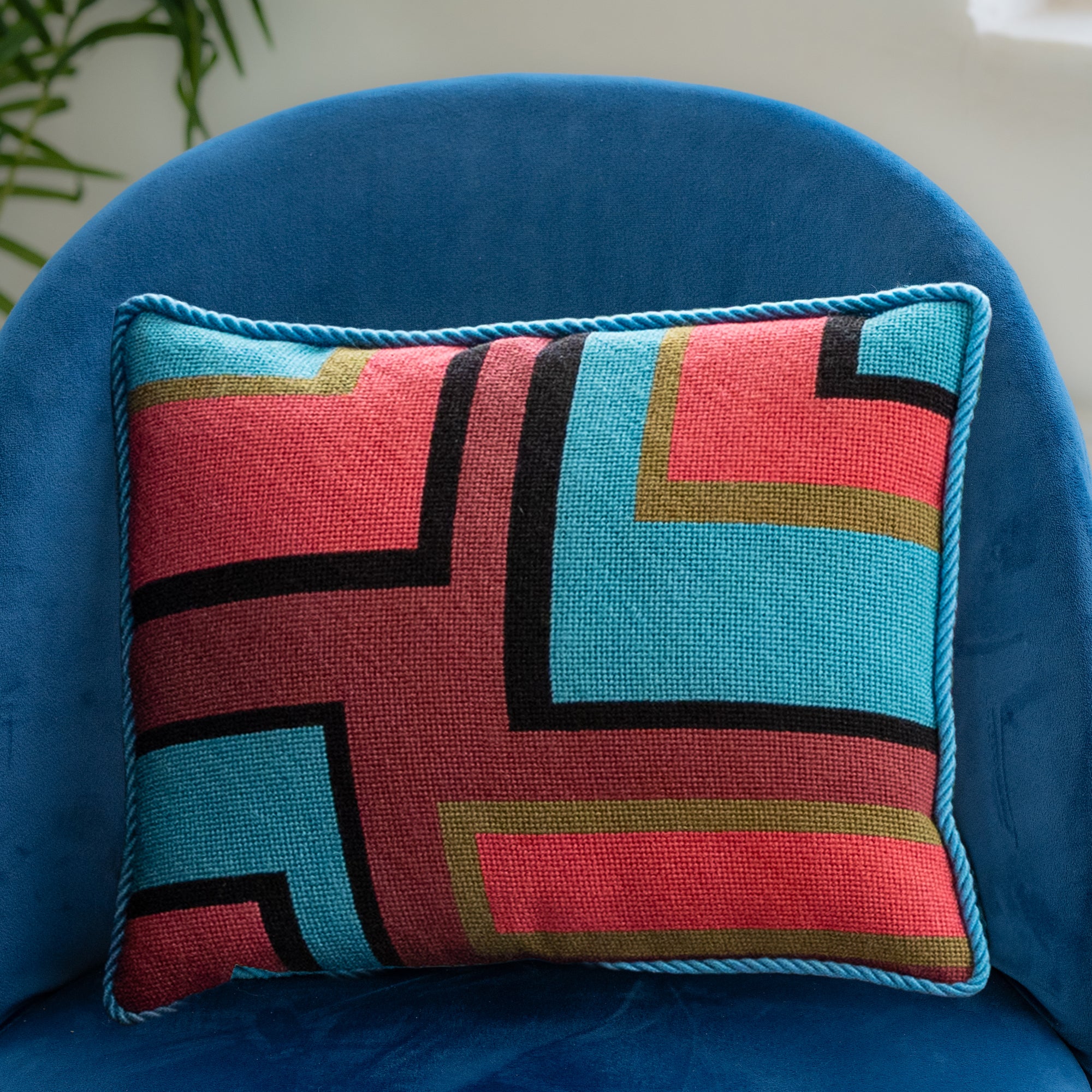 A colorful geometric patterned needlepointed cushion on a blue couch. Designed and sold by Daisy’s Needlepoint Kits.