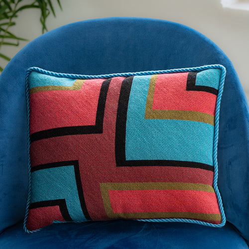 A colorful geometric patterned needlepointed cushion on a blue couch. Designed and sold by Daisy’s Needlepoint Kits.