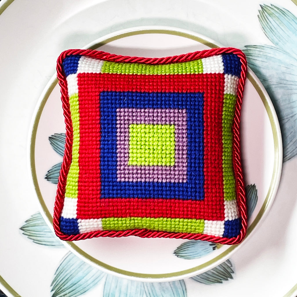A colorful geometric patterned needlepointed cushion on a  small Noritake plate, Up-sa Daisy pattern. Designed and sold by Daisy’s Needlepoint Kits.
