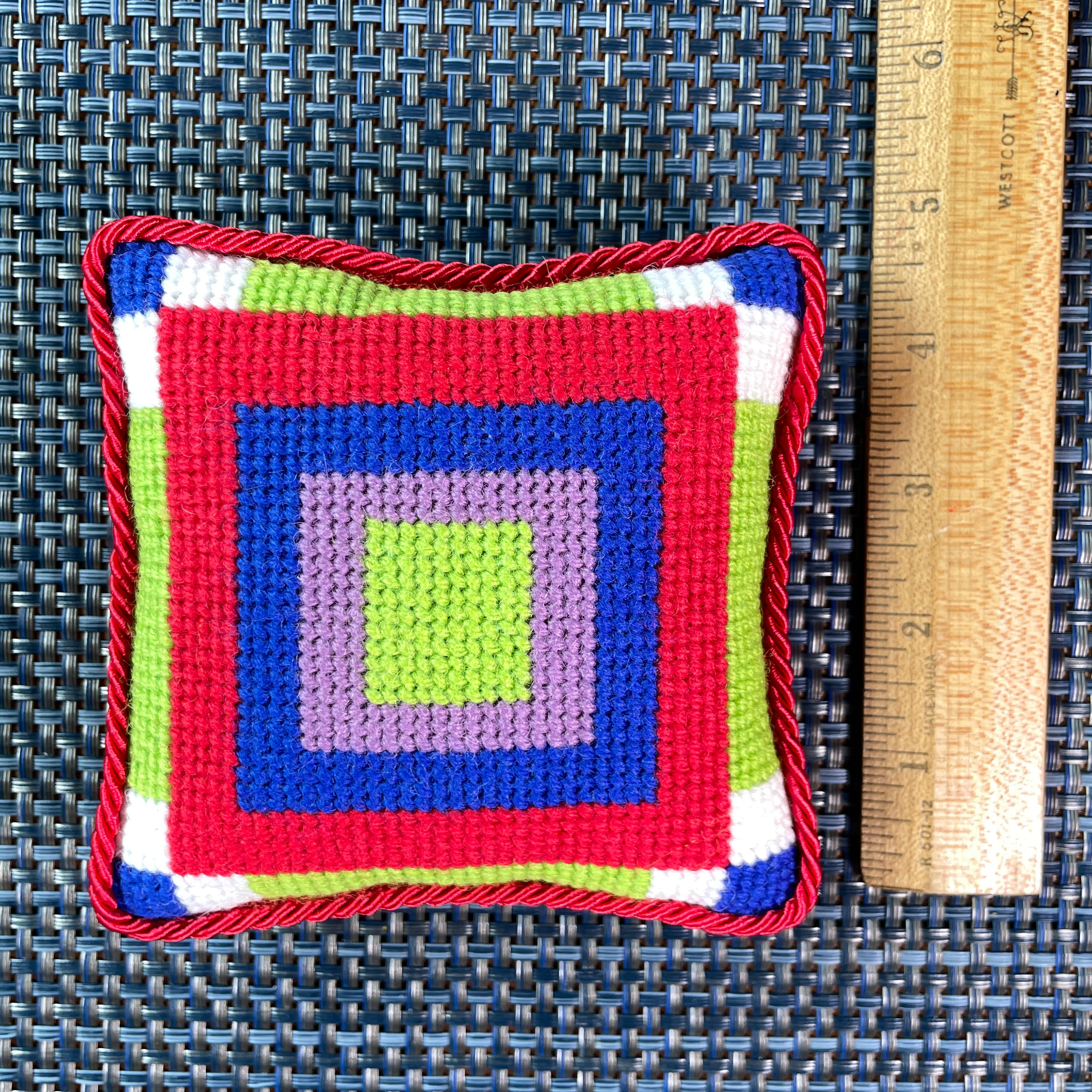 A colorful geometric patterned needlepointed cushion 5 inches by 5 inches, on a blue background. Designed and sold by Daisy’s Needlepoint Kits.

